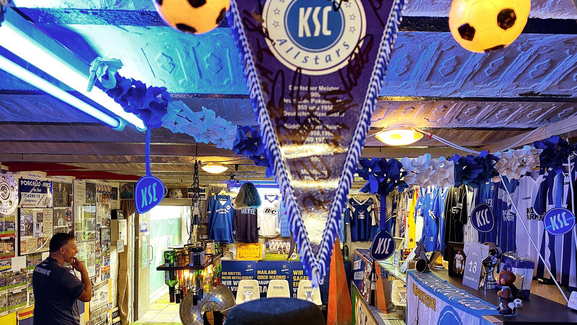 Blaues Licht strahlt durch den Raum des KSC-Museums in Eggenstein. Von der Decke hängen Girlanden und Fahnen. An den Wänden sind Trikots und Poster befestigt.