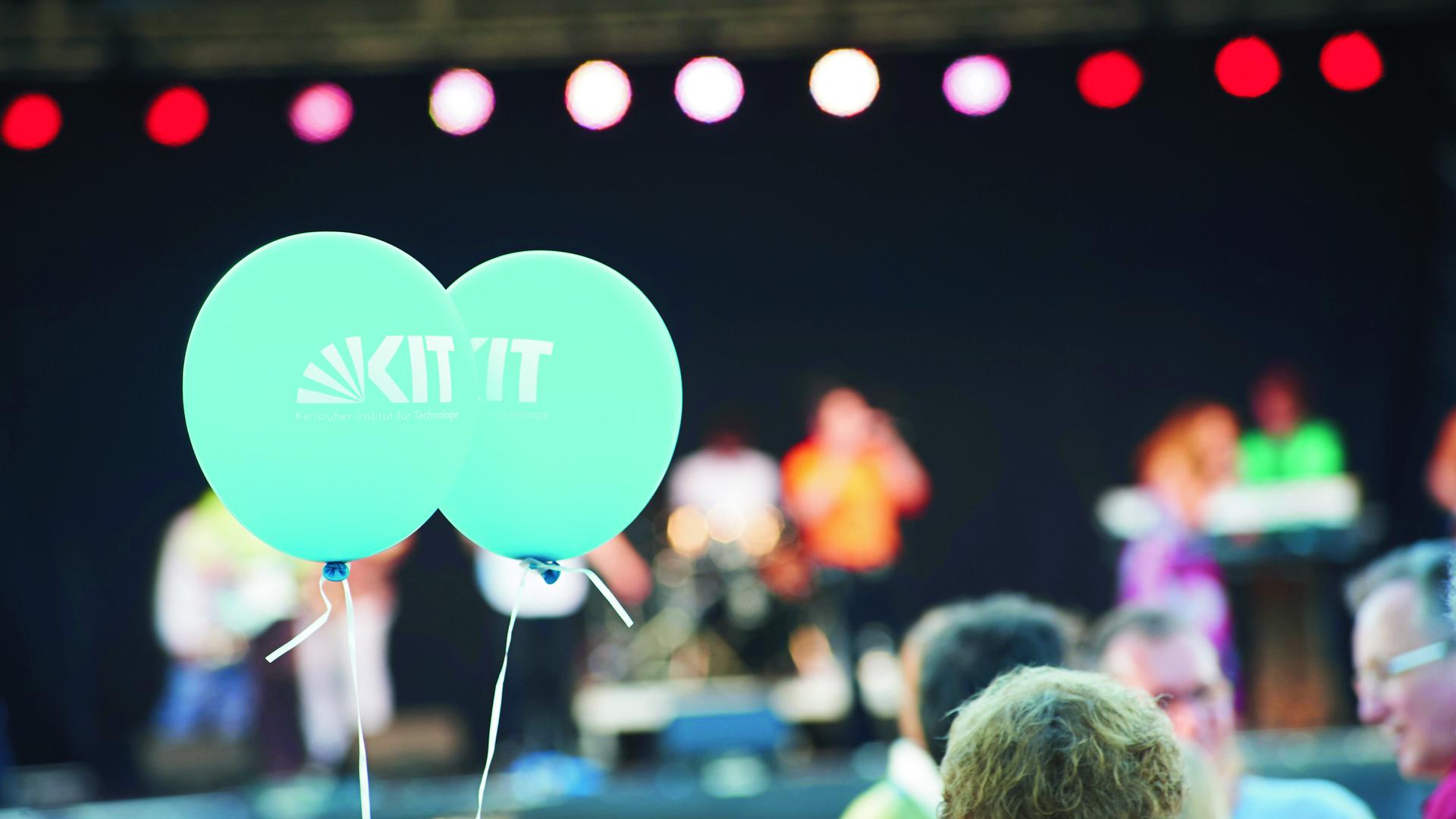 Vor einer Bühne hält jemand zwei blaue Luftballons mit der Aufschrift „KIT“. 