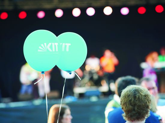 Vor einer Bühne hält jemand zwei blaue Luftballons mit der Aufschrift „KIT“. 