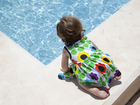 Ein kleines Mädchen in einem bunten Kleid kniet am Beckenrand eines Schwimmbades. 