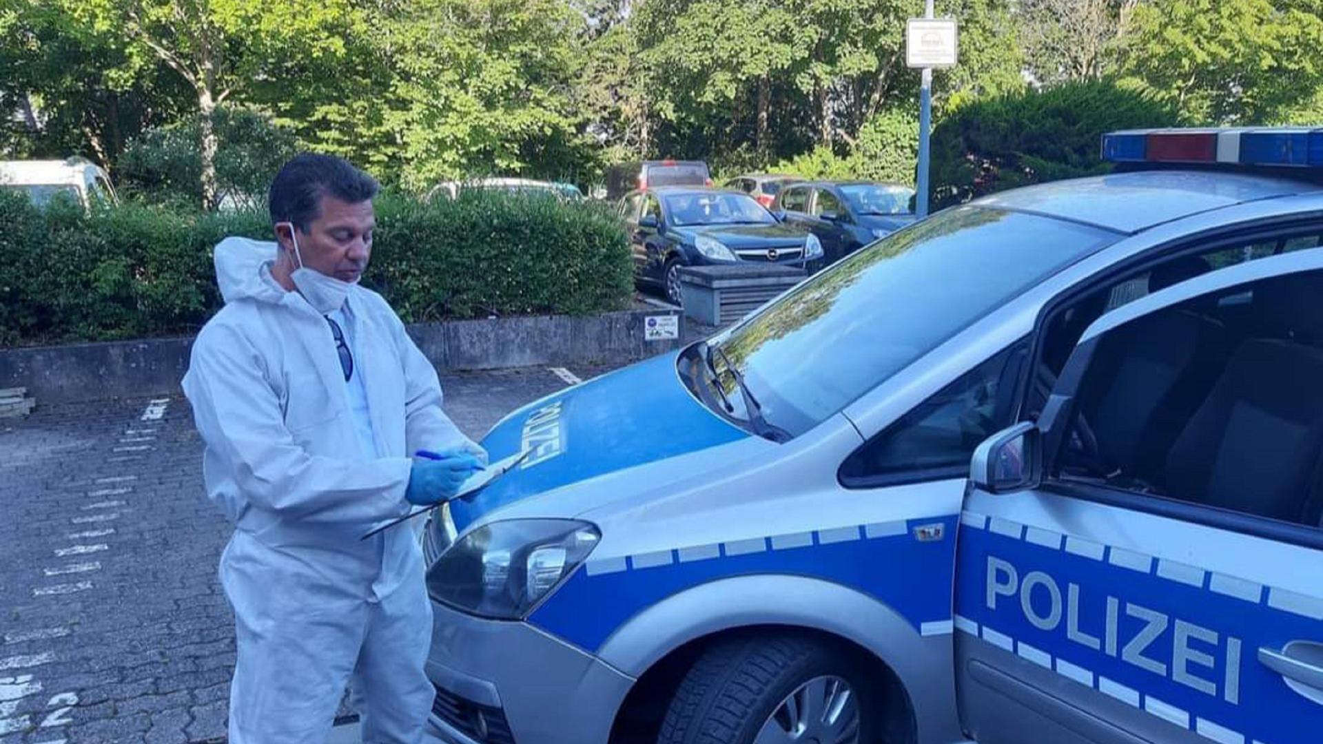 Mann im weißen Overall vermerkt Notizen auf einem Klemmbrett, neben ihm steht ein balues Polizeiauto der Marke Opel. 