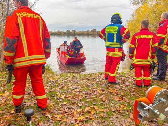 Einsatzkräfte von der Feuerwehr stehen am Ufer eines Sees. Davor ein Boot, das sich vom Ufer wegbewegt. 