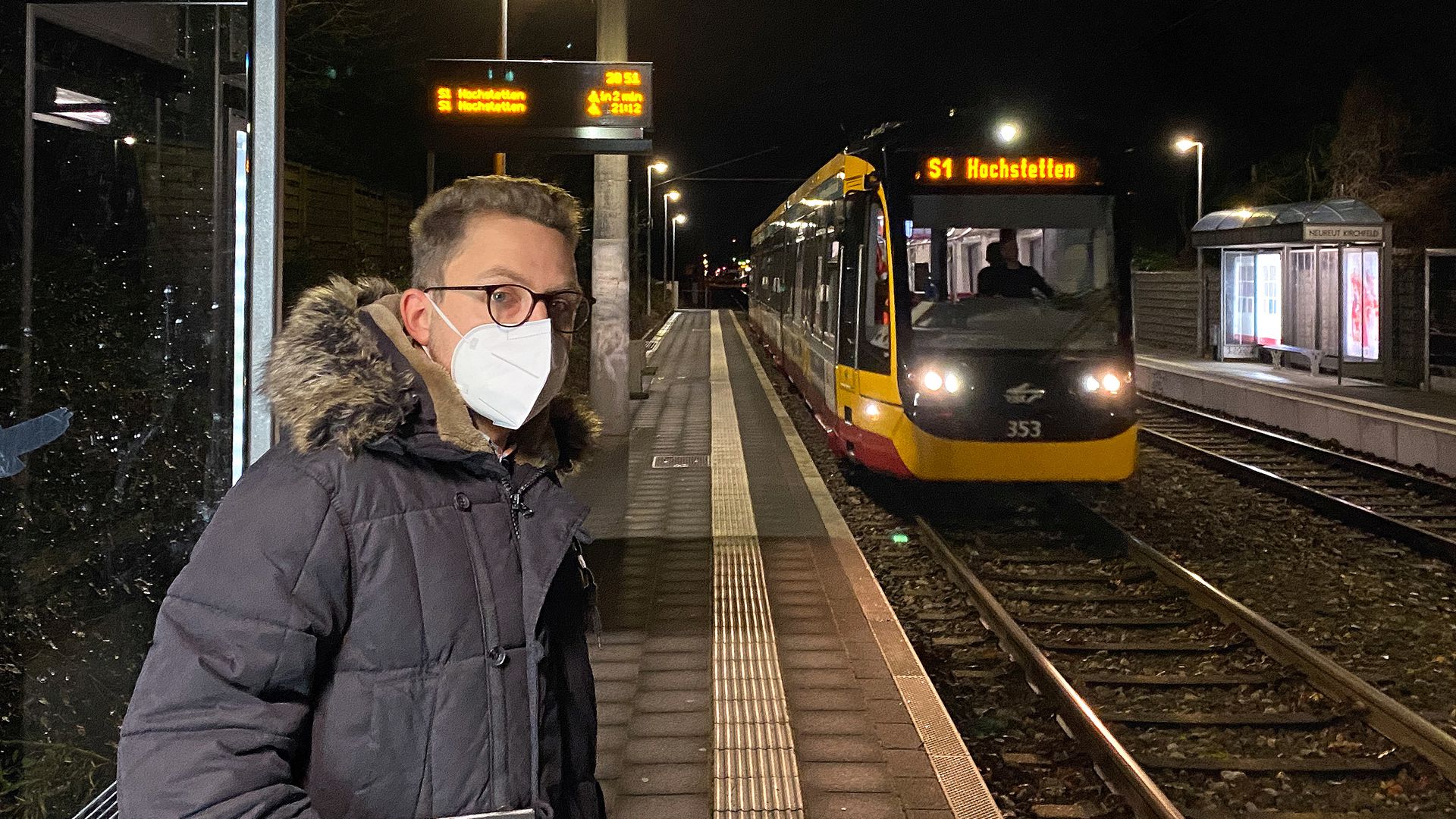 Mann mit Maske nachts am Bahnsteig, im Hintergrund eine Stadtbahn