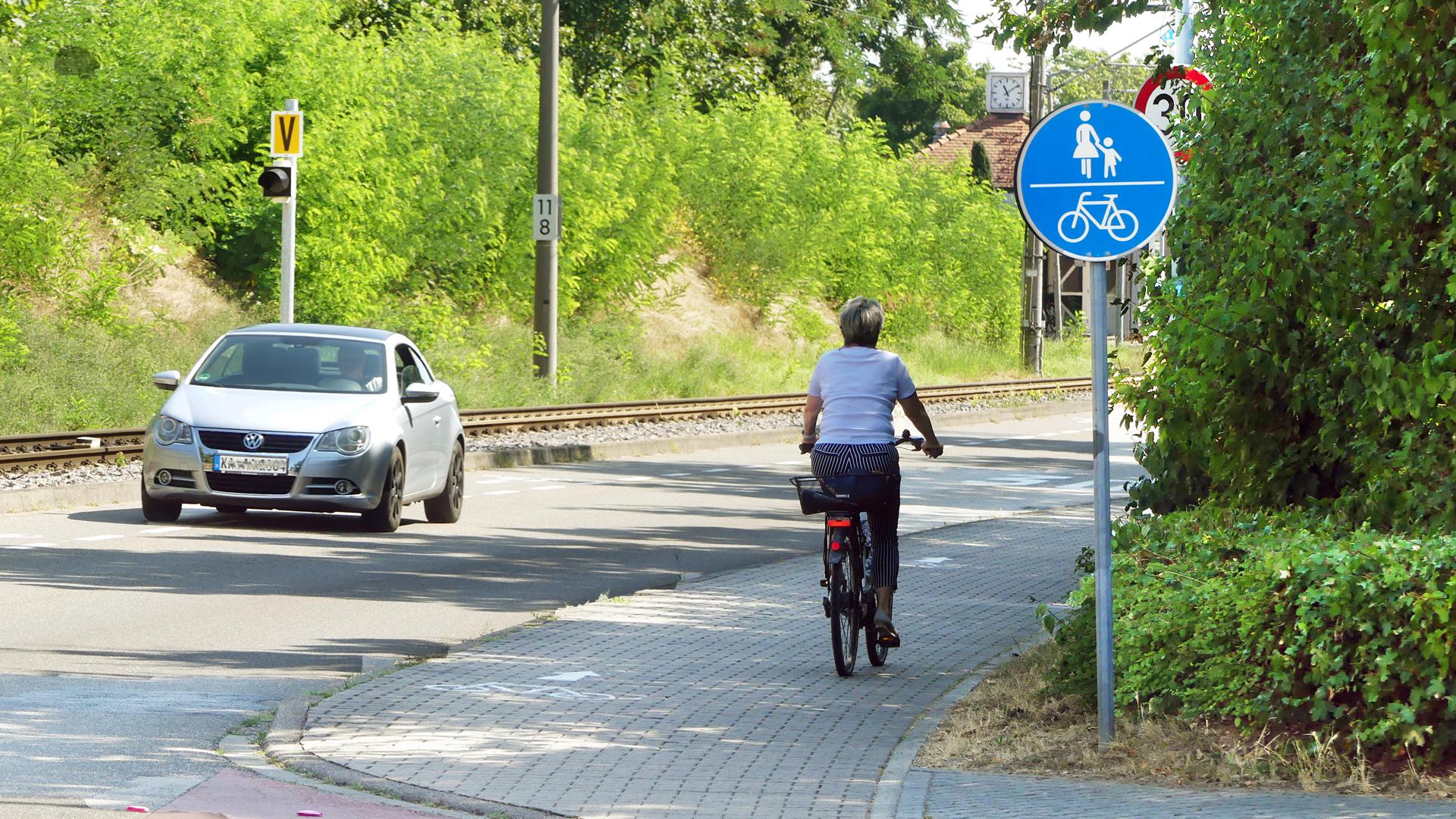 Frau fährt Rad auf Fuß- und Radweg in der Heussstraße in  Linkenheim. Ein Auto fährt auf der Straße.