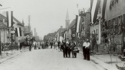 Menschen stehen Anfang der 1930er-Jahre auf der Linkenheimer Hauptstraße, die mit badischen und Hakenkreuzfahnen geschmückt ist. 