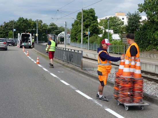 Bauarbeiter sammeln Pylonen von einem Fahrradstreifen ein, daneben fahren Autos vorbei.