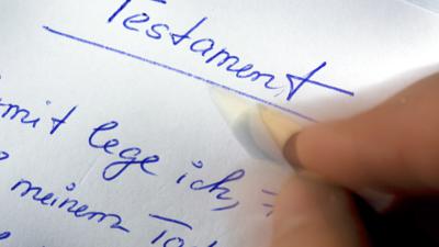 Eine Hand hält einen Stift und schreibt mit blauer Farbe Testament auf ein weißes Blatt Papier. 