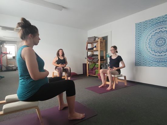 Drei schwangere Frauen sitzen im Stuhlkreis und praktazieren Yoga. 