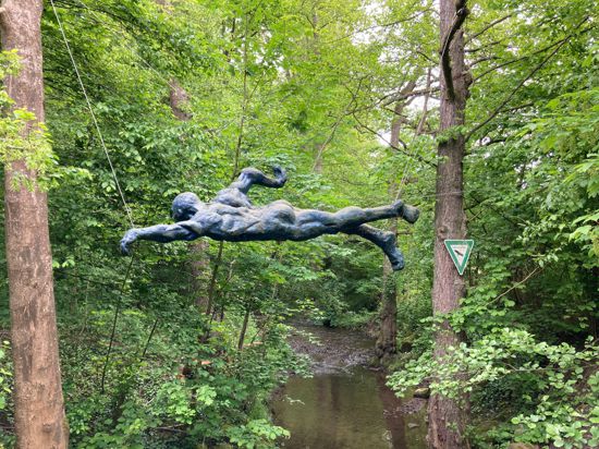 Ein Skulptur, die einen Schwimmer darstellt, hängt an Seilen zwischen Bäumen und schwebt über dem Bocksbach in Kleinsteinbach.