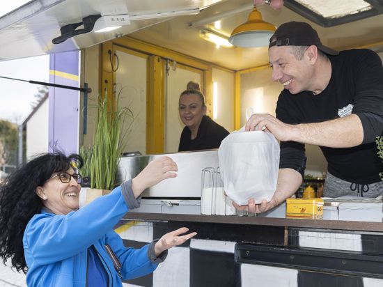 Casim Cilek überreicht Monika Carbone ein Burger-Paket to go, beobachtet von seiner Frau Kader Cilek