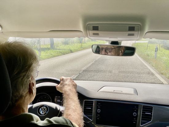 Ein älterer Mann sitzt in einem Auto und hält das Lenkrad fest. 