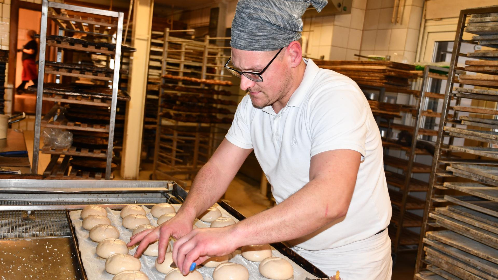Ein Mitarbeiter der Bäckerei Friebolin ritzt gut 20 Brötchen auf einem Backblech mit einem Messer ein.
