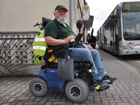 Ein Mann mit seinem Rollstuhl an der Bushaltestelle. Im Hintergrund fährt ein Bus ein.