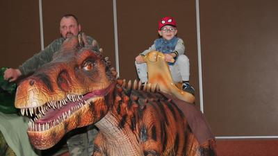Ein Junge sitzt auf einem Plastik-T-Rex, ein Mann steht daneben.