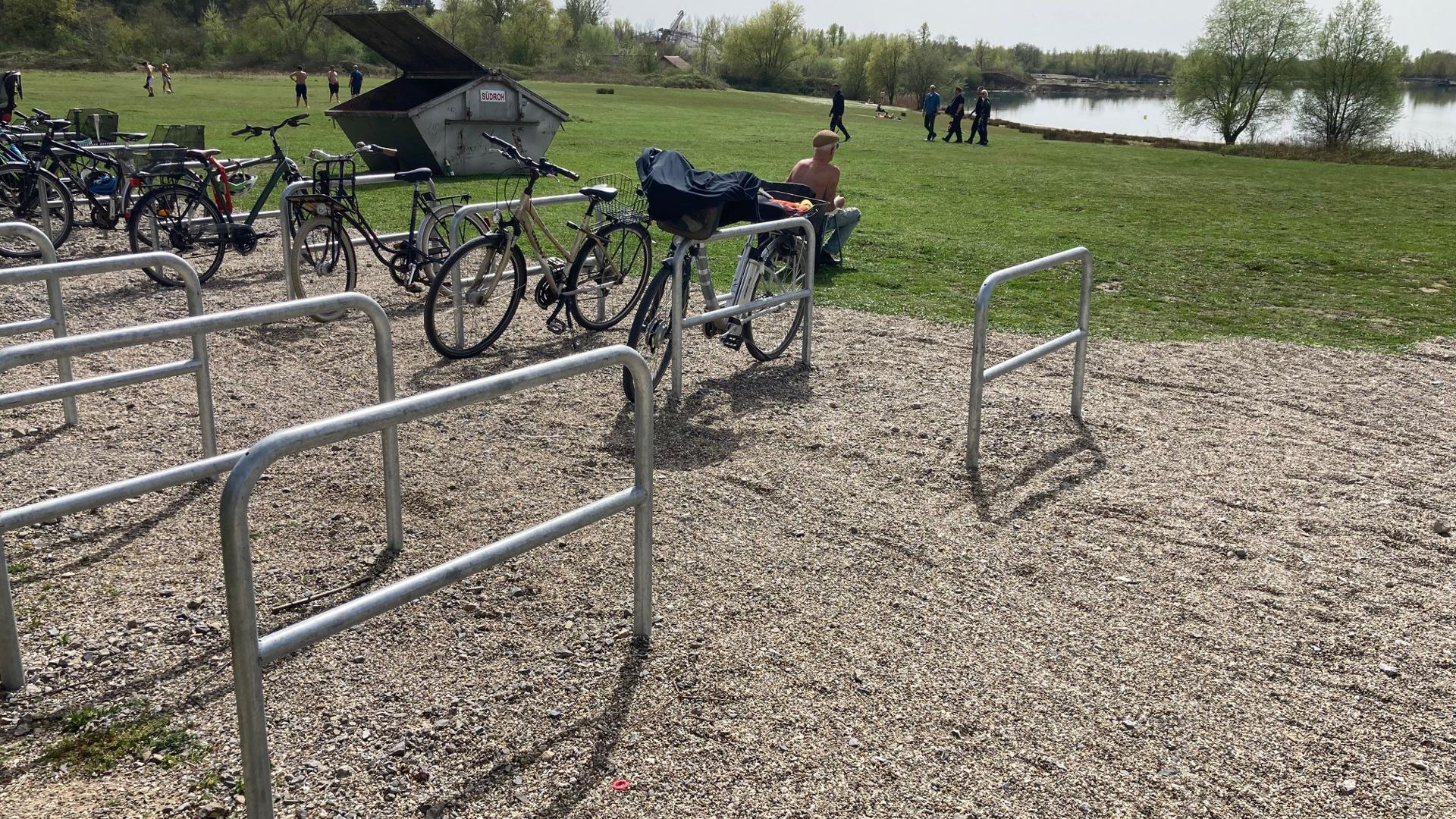 Fahrradbügel stehen am auf einer Schotterfläche, dahinter laufen vereinzelt Menschen über eine grüne Wiese, im Hintergrund ist der Epplesee zu erkennen.