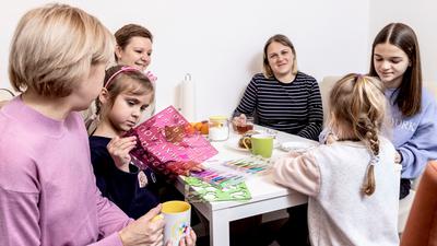 Drei Frauen und drei Mädchen sitzen an einem Küchentisch und unterhalten sich, die Kinder malen und basteln.