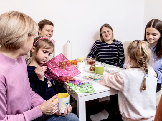 Drei Frauen und drei Mädchen sitzen an einem Küchentisch und unterhalten sich, die Kinder malen und basteln.