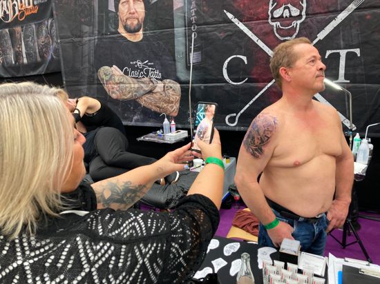 Frau fotografiert einen Mann, der mit noch unfertigem Tattoo posiert