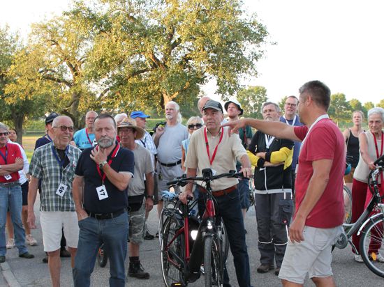 Ein Mann im roten Shirt deutet anderen Männern auf Fahrrädern den Weg. 