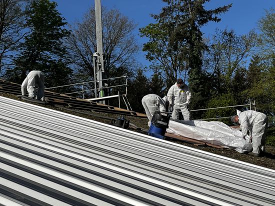 Vier Männer in weißen Schutzanzügen und Atemmasken entfernen asbesthaltige Dachplatten von einem Hof-Dach bei Bretten.