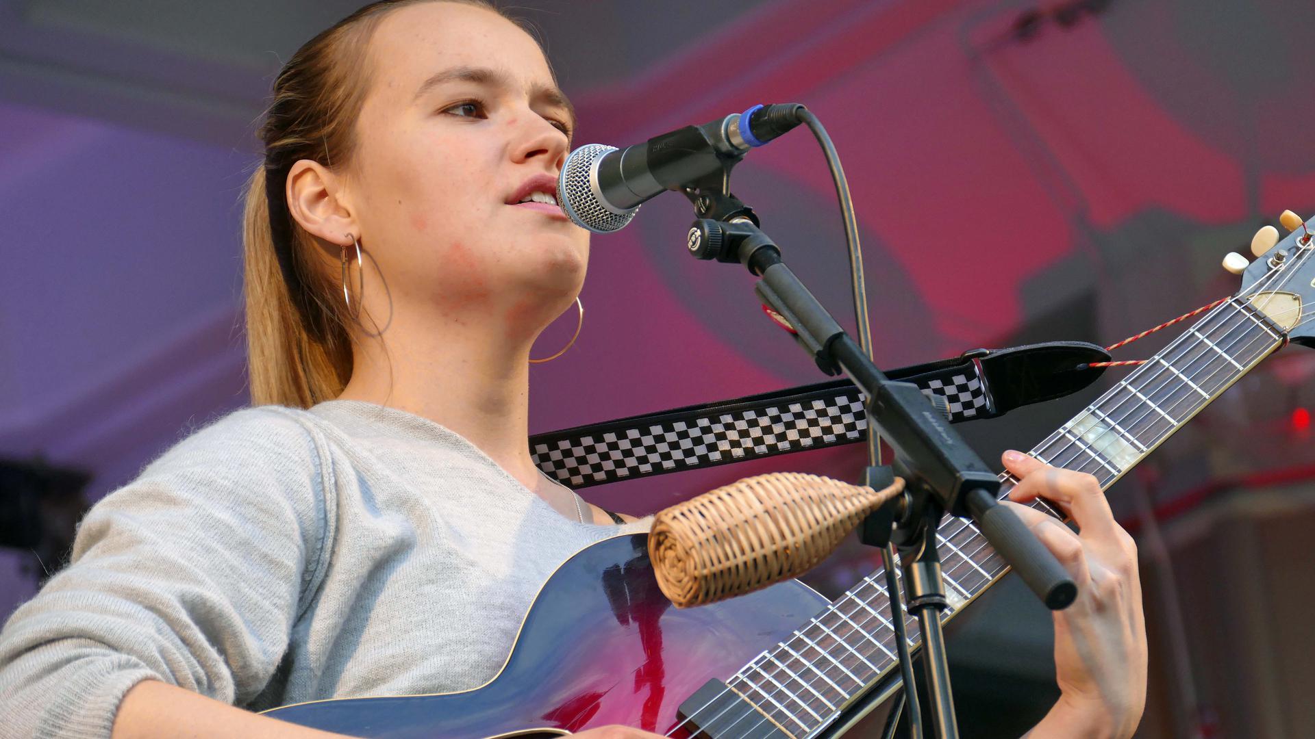 Die junge Musikerin hat ihre blonden Haar zu einem Pferdeschwanz gebunden, spielt Gitarre und singt in ein vor ihr stehendes Mikrofon. 