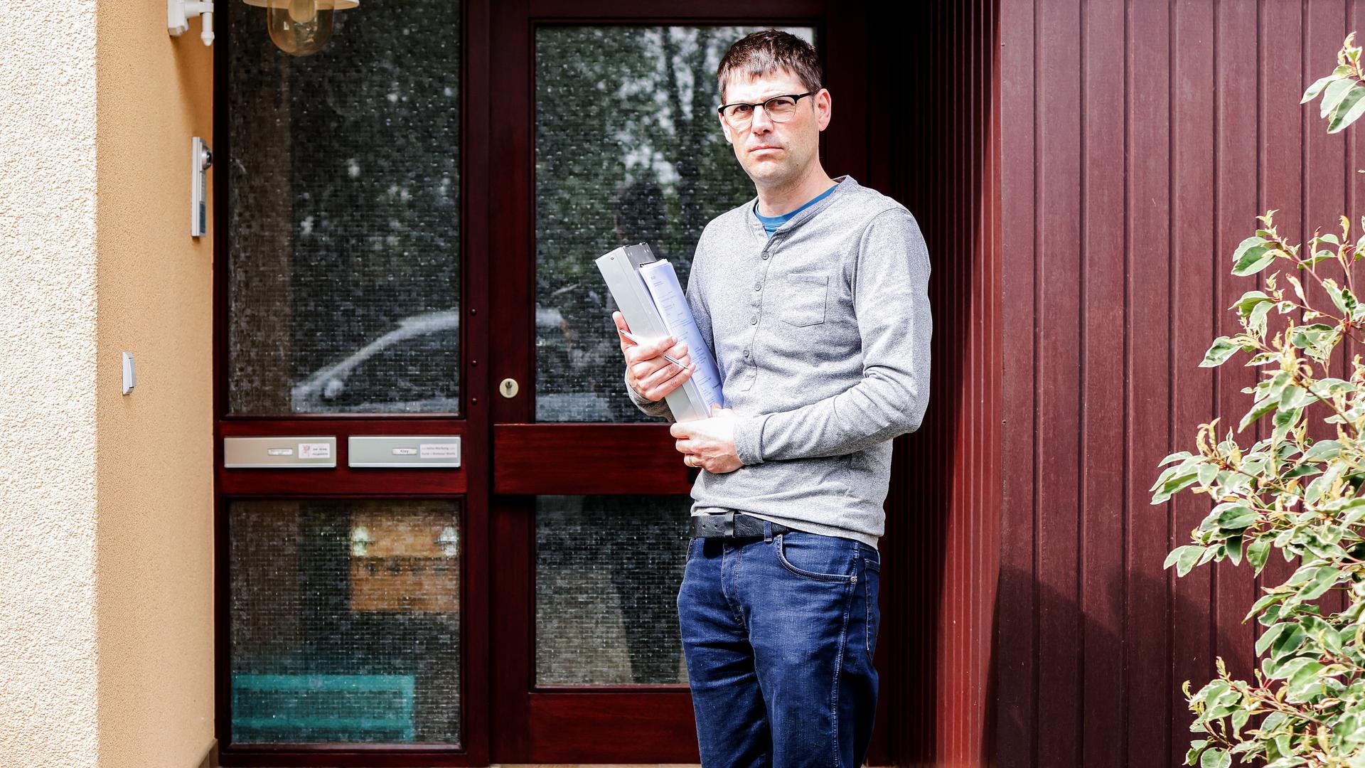Ein Mann mit kurzem schwarzem Haar steht vor einer Haustür und hat ein Aktenpaket im Arm.
