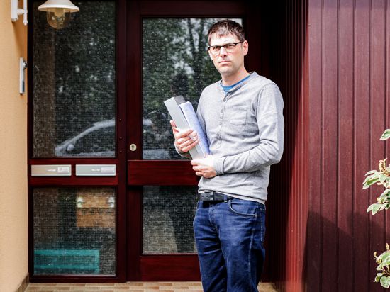 Ein Mann mit kurzem schwarzem Haar steht vor einer Haustür und hat ein Aktenpaket im Arm.
