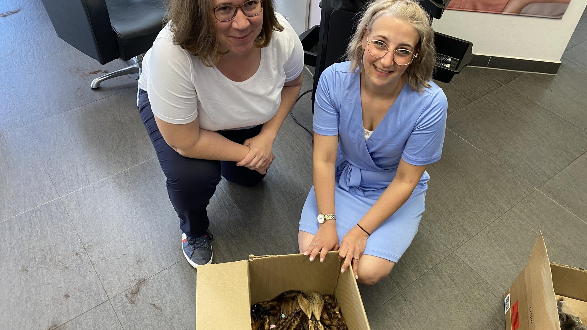 Anna-Lena Nowak und Daniela Schlor knien vor einem Karton. Darin liegen viele Zöpfe aus echtem Haar. Sie gehen als Spenden an Zweithaar-Spezialisten, die Echthaar-Perücken daraus fertigen.