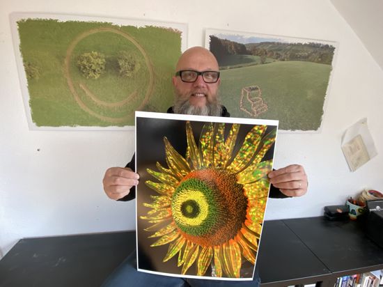 Jürgen Scheible aus Oberderdingen ist ein digitaler Künstler. Im Hintergrund 2 Fotos seiner „dronelandart“, bei der er Gegenstände installiert und von oben mit der Drohne an einem speziellen Punkt so filmt, dass 3-D-Effekte entstehen. In den Händen hält er ein Foto einer Sonnenblume, die er mit Hilfe seiner Lichtkunst und seiner Software „MobiSpray“ mit dem Handy und einem Projektor in neue Farben getaucht hat.