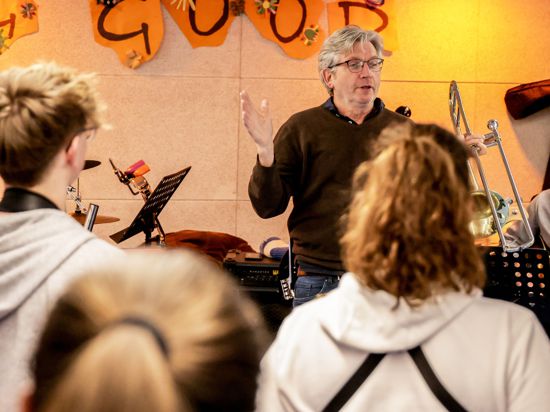 Ein Mann mittleren Alters steht zwischen Schülerinnen und Schülern bei der Probe der Big Band des Melanchthon Gymnasiums. Sie haben Saxofone, Trompeten und andere Instrumente in ihren Händen.