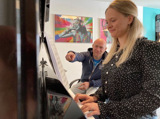 Eine junge blonde Frau sitzt am Klavier und im Hintergrund ihr Musiklehrer. 