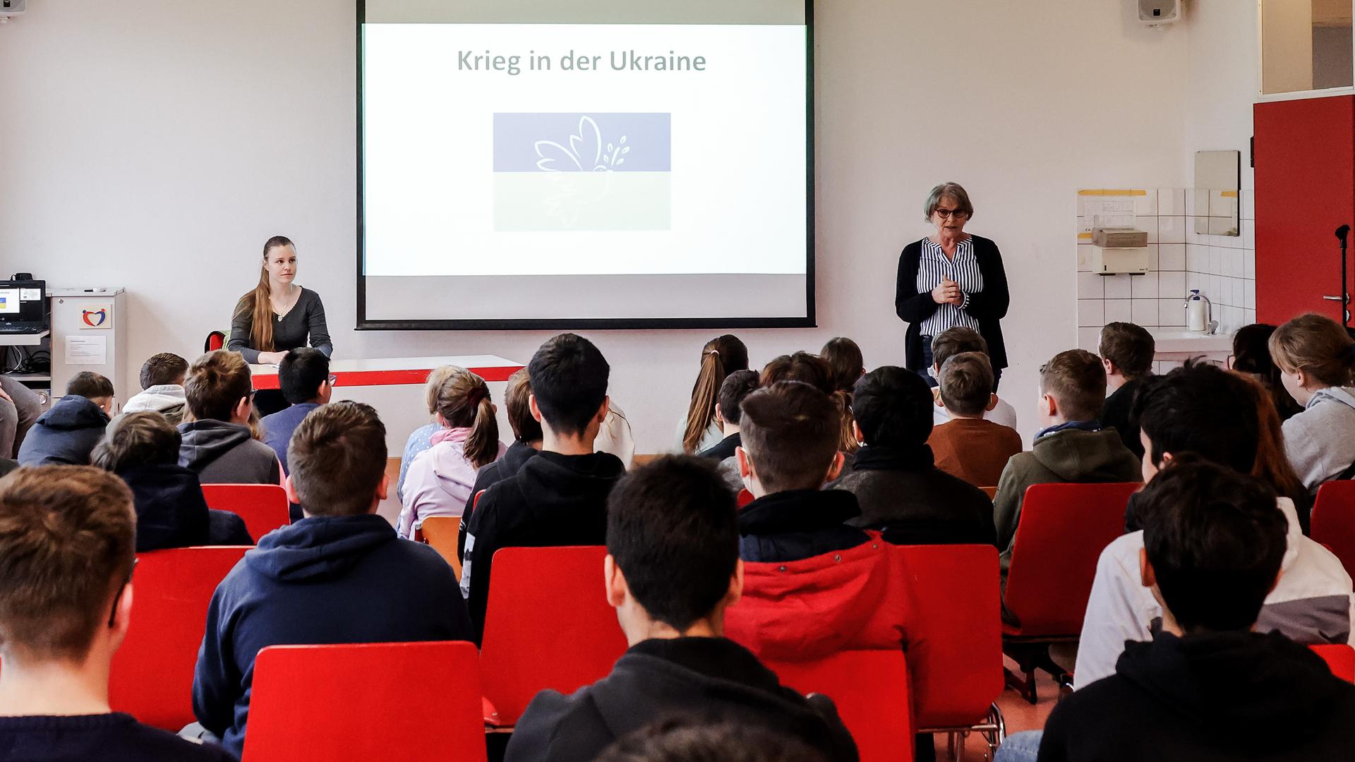 Zwei Frauen sehen sich in einem großen Klassenraum rund 30 Schülerinnen und Schülern gegenüber. In ihrer Mitte steht auf einem Whiteboard „Krieg in der Ukraine“. 