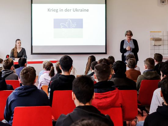 Zwei Frauen sehen sich in einem großen Klassenraum rund 30 Schülerinnen und Schülern gegenüber. In ihrer Mitte steht auf einem Whiteboard „Krieg in der Ukraine“. 