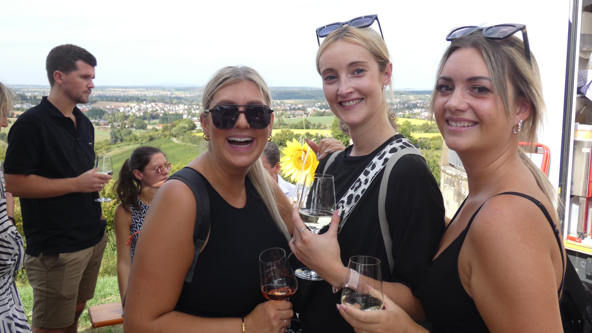 Drei Frauen in Partykleidung halten Weingläser in der Hand. Im Hintergrund sind weitere Personen zu sehen und ein Ausblick über eine Landschaft.