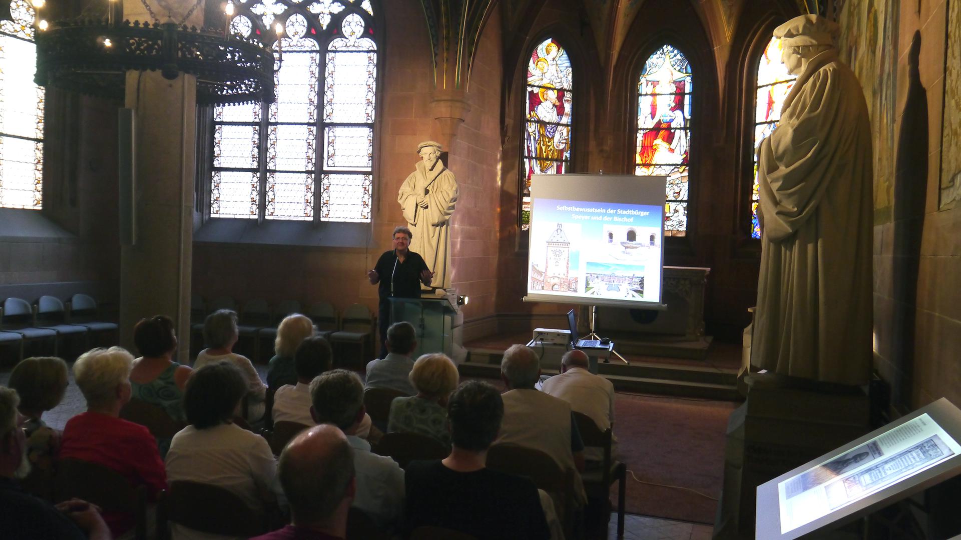 Eine beleuchtete Leinwand steht in einem Kirchengebäude. Ein Mann spricht zu einem Publikum, das von hinten zu sehen ist.