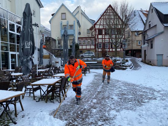 Zwei Männer in der signal-orangenen Arbeitskleidung des Bauhofs Bretten schippen Schnee und Streuen den Weg im Löwenhof.