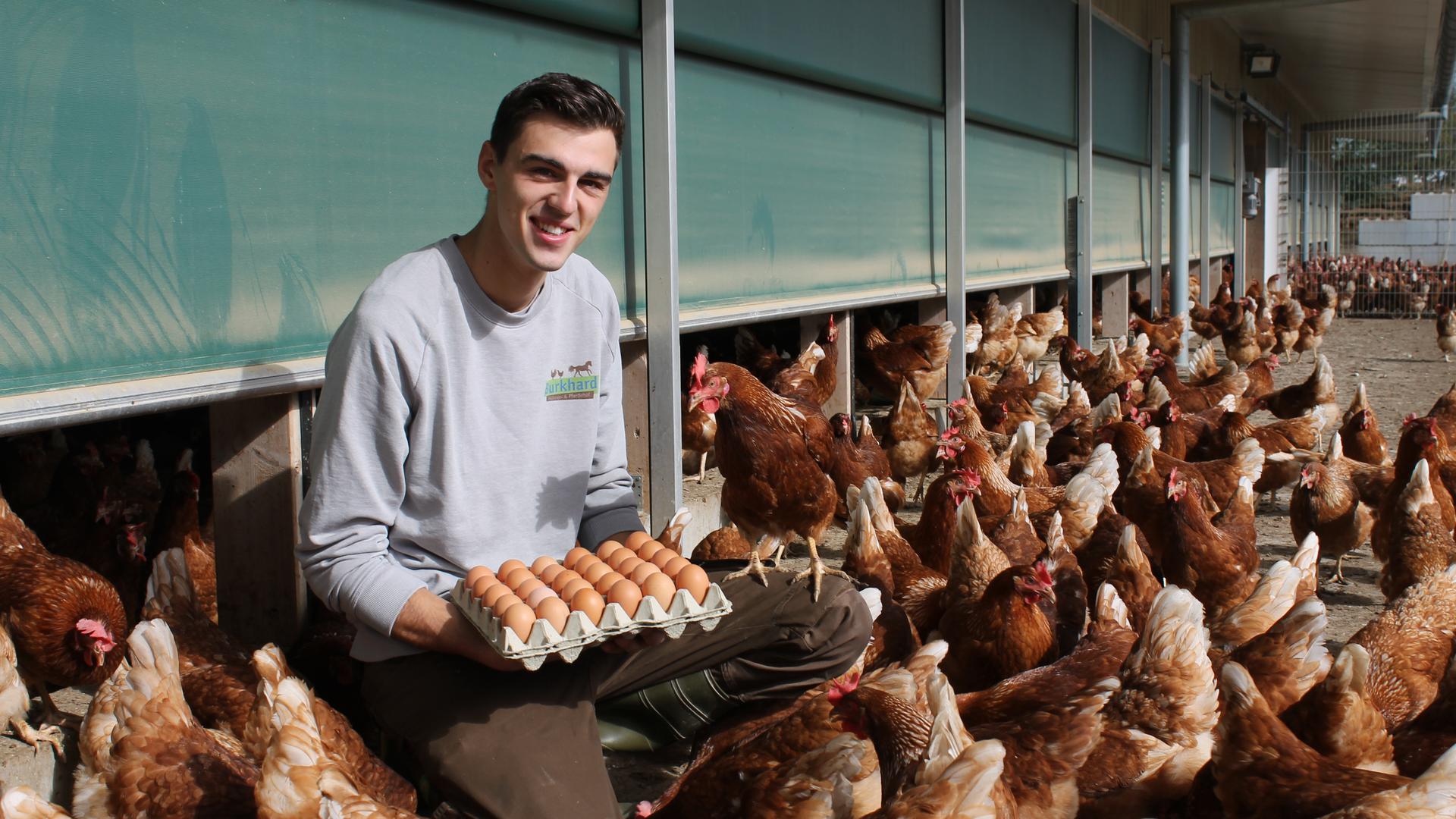 Tobias Burkhard kniet inmitten seiner Hühner und präsentiert frische Eier.
