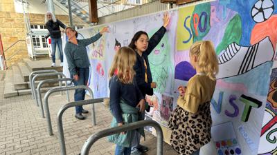 Zusammen mit Schülern rollen Künstler Tom Rebel und Fachschaftsleiterin Anne Kempter die Banner vor der grauen Betonwand auf dem Schulhof aus.