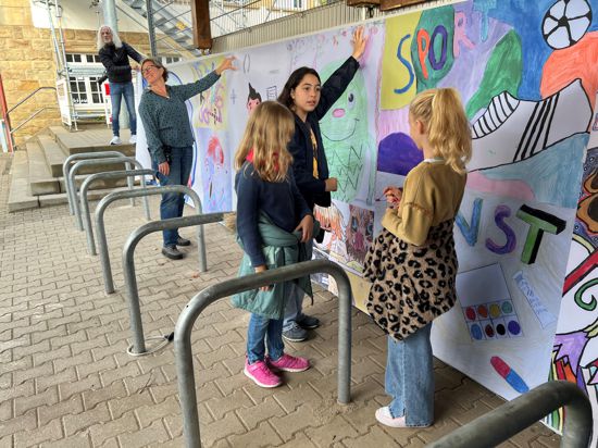 Zusammen mit Schülern rollen Künstler Tom Rebel und Fachschaftsleiterin Anne Kempter die Banner vor der grauen Betonwand auf dem Schulhof aus.