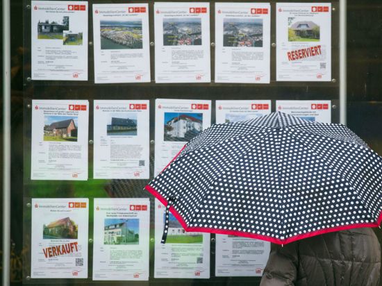 Auf diesem Bild steht eine Frau mit Regenschirm vor einem Schaufenster mit Immobilienangeboten.