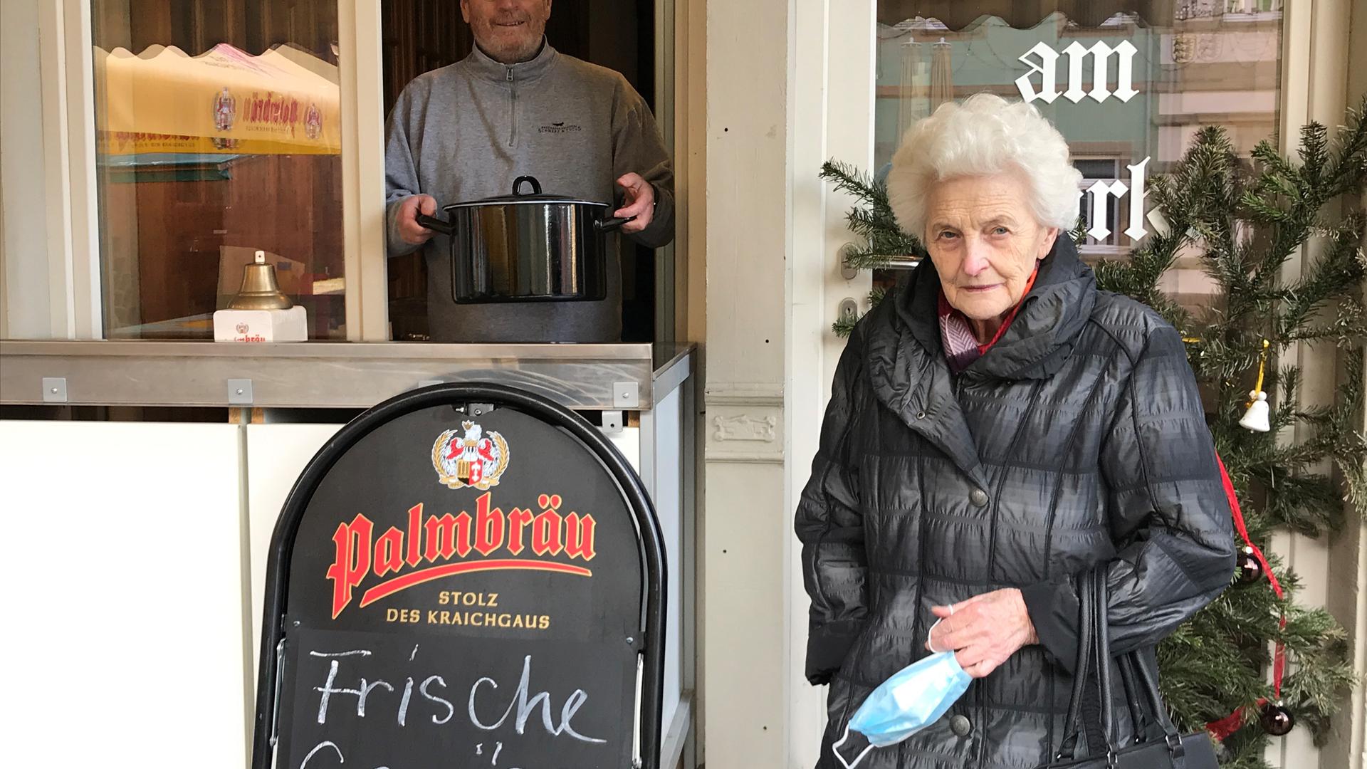 Vor dem geöffneten Fenster eines Kiosks steht eine Seniorin. Im Fenster lächelt ein Mann. Er hält einen Topf in seinen Händen. 