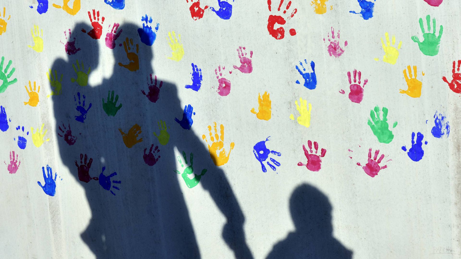 Kindergärten sind ein Kostenfaktor in Bruchsal. Auf dem Bild ist ein Mann  zu sehen, der mit einem Kind auf dem Arm und einem an der Hand einen Schatten auf eine mit bunten Handabdrücken bemalte Wand einer Kindertagesstätte wirft. 