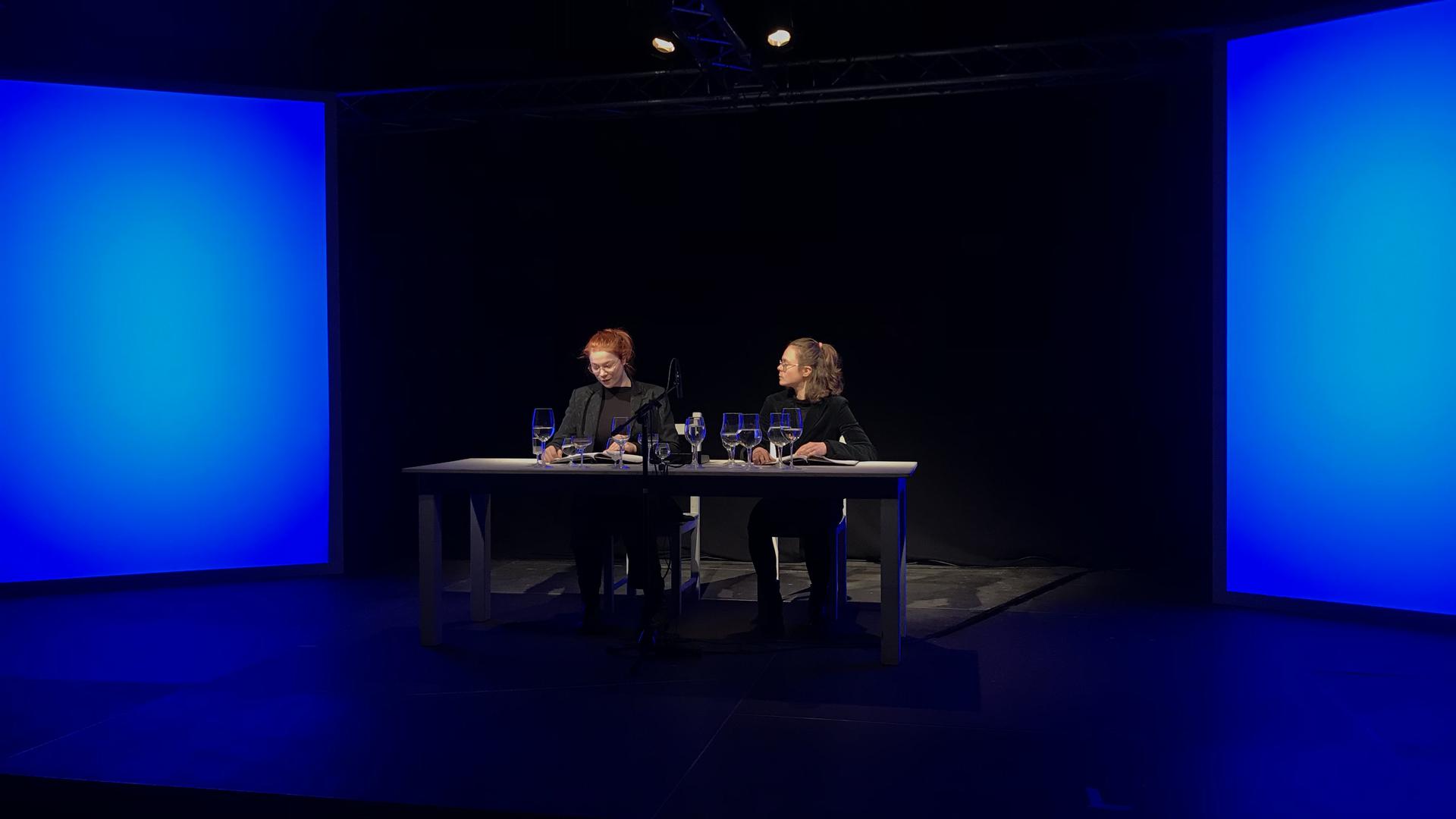 zwei Frauen sitzen an Tisch auf Bühne in blauem Licht