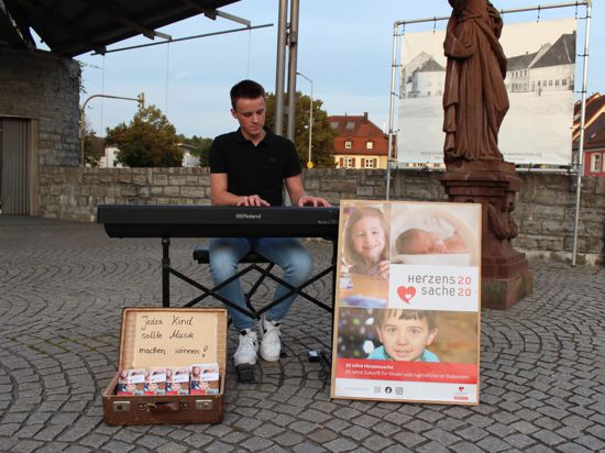 Ein junger Mann, Laurin Sigmund, sitzt in Bruchsal auf dem Fußgängerweg an seinem Piano und spielt. Davor stehen Plakate und ein Spendenkoffer für eine Hilfsaktion.