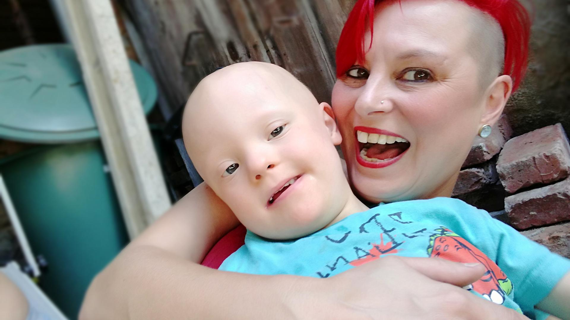 Eine Frau mit knallroten Haaren hat ein Kind im Arm und lacht in die Kamera. 