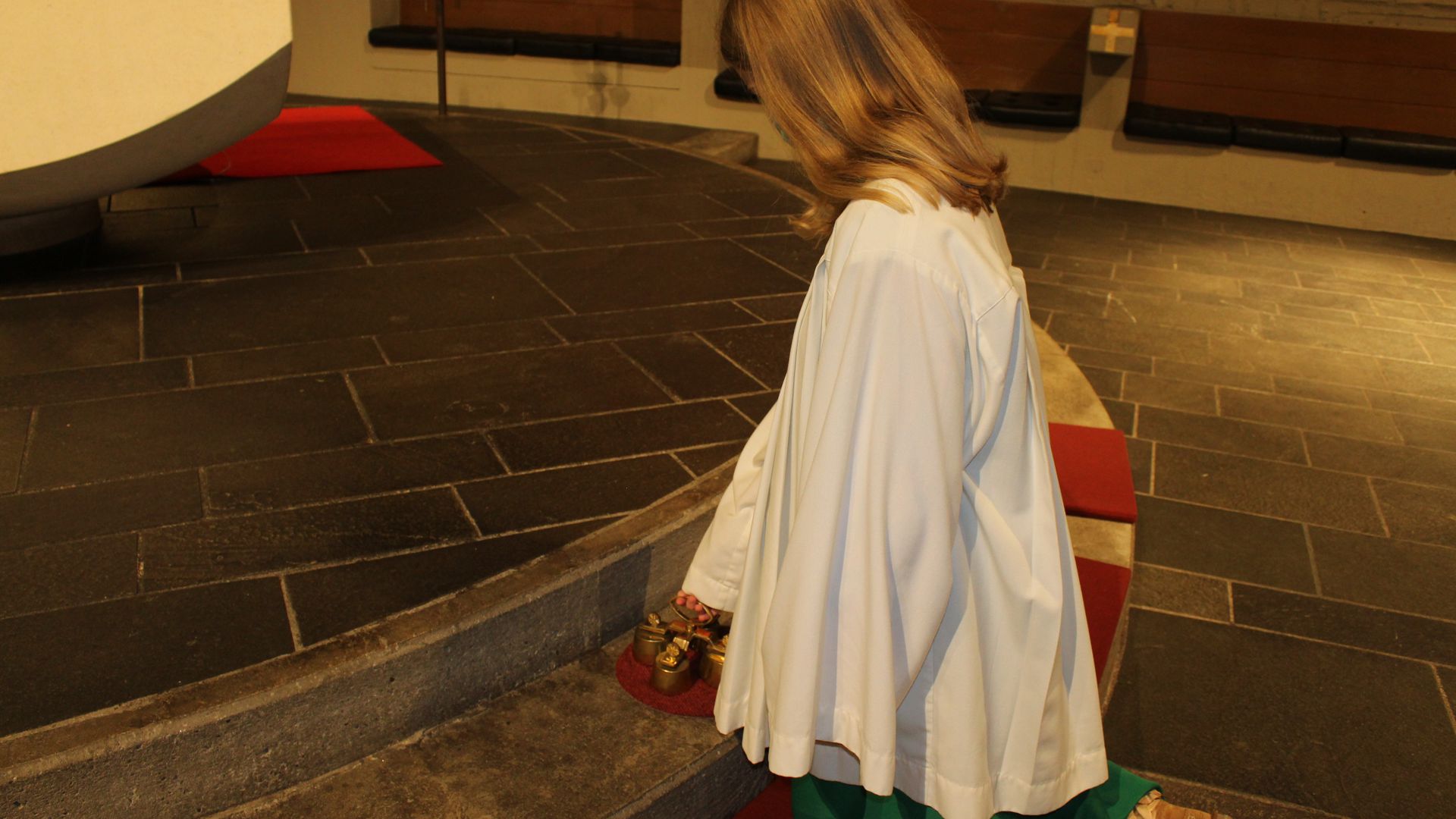 weibliche Ministrantin mit grünem Unterrock kniet vor dem Altar und hat die Glöckchen vor sich