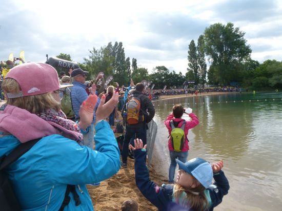 Hunderte Menschen stehen an einem Baggersee-Strand, einige halten Kameras oder klatschen in die Hände.