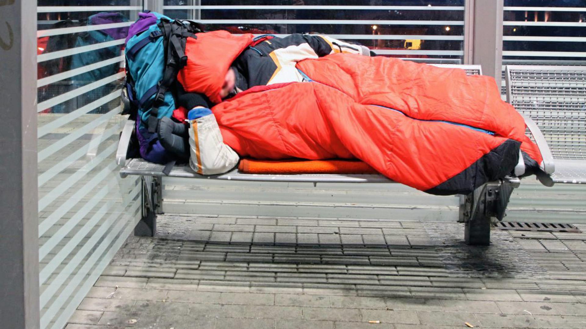 Eine obdachlose Person schläft in einem Schlafsack auf einer öffentlichen Sitzgelegenheit.