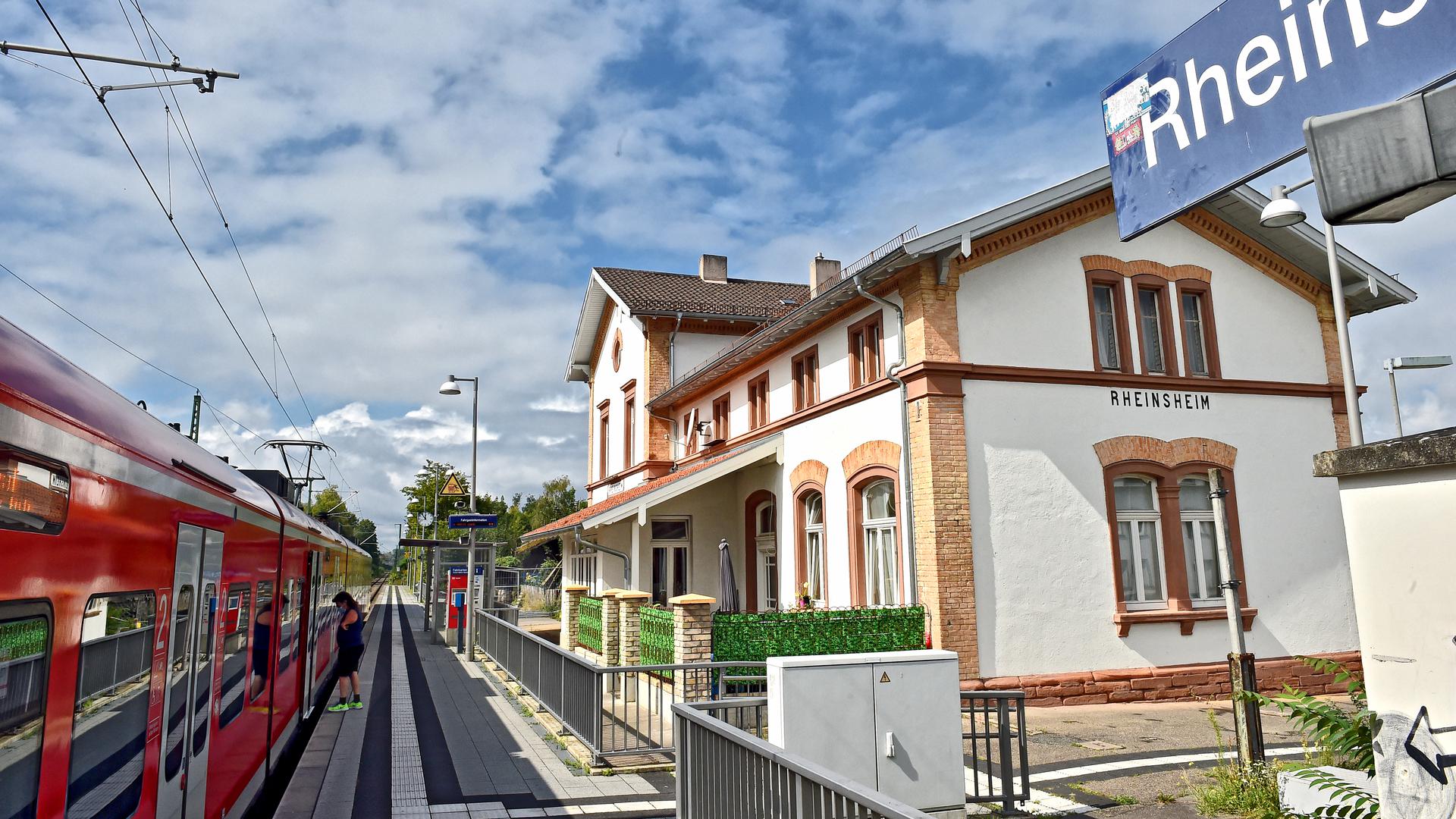 Blick auf den Bahnsteig in Rheinsheim: Auf der rechten Seite steht das weiß-braune Bahnhofsgebäude. auf dem Bahngleis auf der linken Seite fährt ein roter Zug ein. 