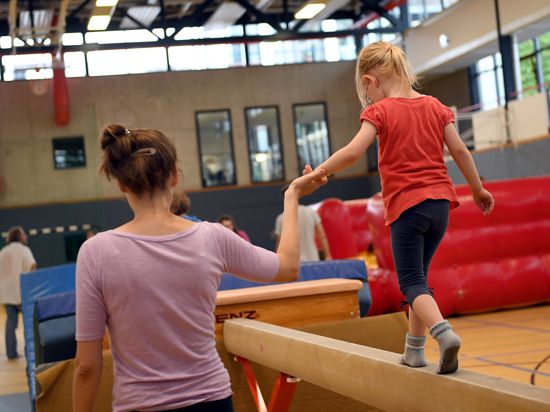 Eine Frau hält ein Kind an der Hand, das in einer Sporthalle über einen Schwebebalken balanciert.